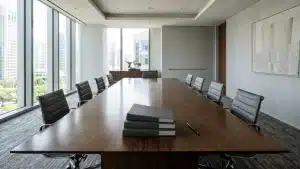 A modern corporate meeting room featuring a large dark wood table in the center, surrounded by gray office chairs. On the table, there is a stack of folders and a pen. In the background, large glass windows offer a view of urban buildings.